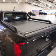 A black VW Amarok pickup truck parked inside a showroom, featuring a black slatted tonneau cover on the truck bed.